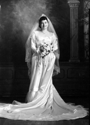 Studio wedding portrait of María Oralia "Orie" Durán Trujillo of Denver, Colorado. She wears a long, silk wedding gown with a scoop neck, trimmed in lace, a veil trimmed in lace and holds a bouquet of flowers. María married Filbert G. Trujillo, sang in the Saint Cajetan's church Mariachi Alegre de San Cayetano, and directed the Southwest Hispanic Colonial Dancers.