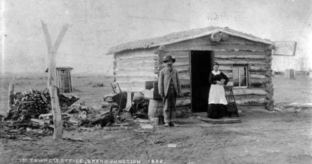 View of Grand Junction Town & Development Company's first office, on 2nd (Second) and Ute Streets, in Grand Junction, Colorado, with R.D. Mobley and Mrs. Jackson posed in front of the small one-room log cabin with sod roof. A "dress making" sign is attached to the cabin and a pile of firewood is between two posts supporting wire or string. Outhouses in back of the cabin show. The cabin is thought to be the first structure built in Grand Junction with R.D. Mobley the first postmaster.