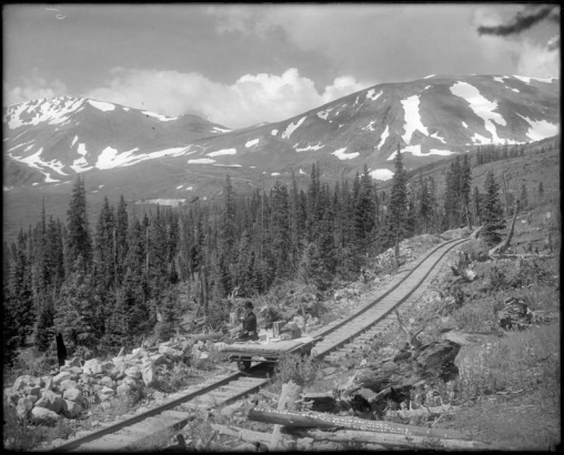 View of Argentine Central Railway narrow gauge track with push car near timberline, Argentine Pass, Clear Creek County, Colorado. Waldorf Mine distant center; Rocky Mountains with snowfields; man on push car with derby hat.