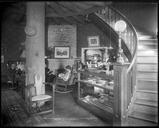 Hotel Glenisle, office area, Glenisle, Colorado, reached via Colorado and Southern Railway; interior view, man standing behind display case containing cigars, fishing poles and supplies with ledger on top; man and women reading magazines seated in rocking chairs in front of brick and stone fireplace; massive log center support, persian or oriental rug with spittoon; entryway into dining room behind display case.