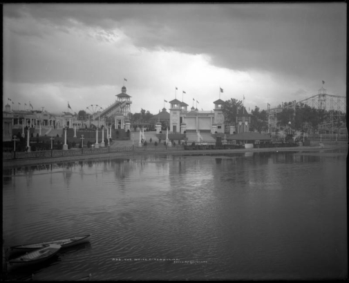 View across Lake Rhoda towards the "White City" (later called Lakeside Amusement Park), Lakeside, Colorado, near Denver; shows the Big Splash ride, ferris wheel, Scenic Railway depot and two miniature steam locomotive engines and cars (train), roller coaster, clock, boats in lake, and rain clouds.