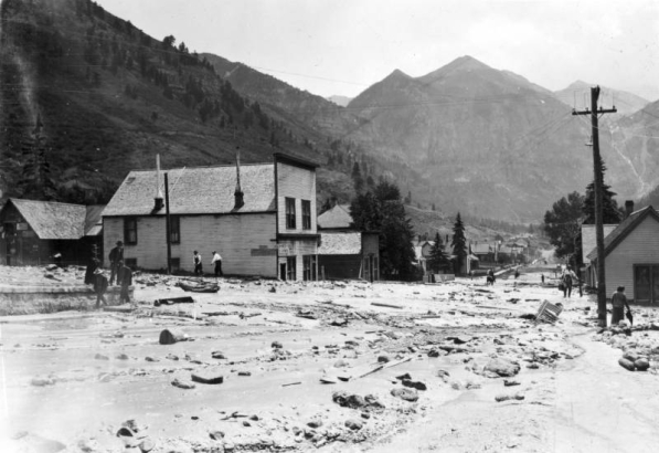 View east down mud and debris-filled street from the disastrous Cornet Creek flood on July 27, 1914, Telluride, Colorado; shows two-story false front, Iowa House on corner, other wood frame businesses, and residences. Men and woman make their way along streets on foot and horseback. San Juans and Ingram Falls are in background.