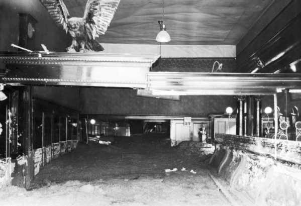 Interior of Sheridan Hotel Bar with mud and high-water marks from  the disastrous Cornet Creek flood on July 27, 1914, Telluride, Colorado. Calendar on back wall marks date; room includes a clock high on left wall, stuffed bird with outstretched wings on top decorative molding, bar, cash register, and electric lights.