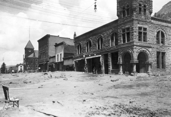 View northwest down Colorado Avenue after the Cornet Creek flood on July 27, 1914, Telluride, Colorado; shows First National Bank Building on corner of Fir and Colorado, three false front businesses, the Sheridan Hotel, the San Miguel County Courthouse, and mud and debris- filled street. Townspeople are near the bank and in distance in front of the Sheridan with a horse and automobile; electric and telephones lines cross street; chairs and tables are part of debris.