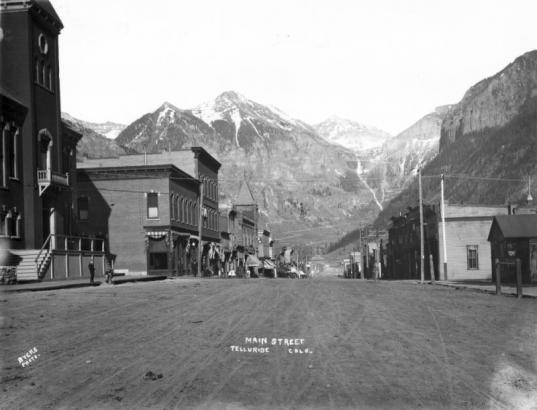 View east down Main Street (Colorado Avenue) Telluride (San Miguel County), Colorado. Ajax, Telluride and Ingram Peaks with light snow and Ingram Falls are in background. Buildings include the San Miguel County Courthouse, the Sheridan Hotel; First National Bank, and the Telluride Mercantile Company block.