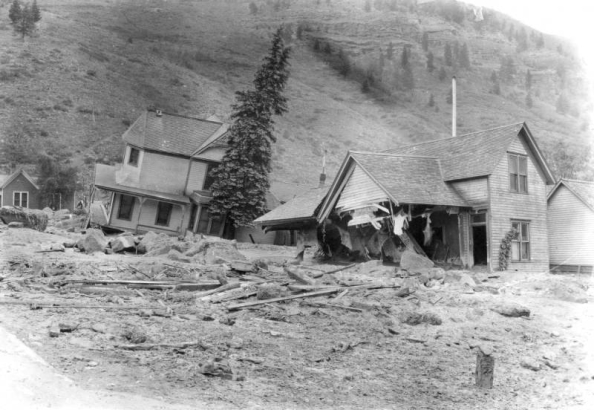 Oak Street filled with mud and debris from the disastrous Cornet Creek flood on July 27, 1914, Telluride, Colorado. Jack Hawkins' two-story wood frame Victorian with front wraparound porch and second-story bay window tilts on side from flood waters; smaller wood frame structure with front intersecting gables destroyed in foreground; cliffs in background.