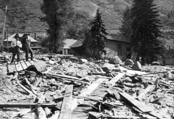 Two men stand on top of debris brought down by the Cornet Creek flood on July 27, 1914, Telluride, Colorado. Mud and debris fill street with damaged wood frame structures; Telluride Hospital is in background with building leased to San Miguel County Historical Society for use as a museum.
