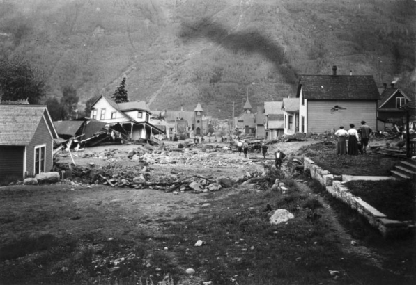View south down Oak Street filled with mud and debris from the disastrous Cornet Creek flood on July 27, 1914, Telluride, Colorado. Townspeople survey damage. Jack Hawkins' two-story wood frame Victorian with front porch and second story bay window tilts on side from flood waters; smaller destroyed structure is next door; residences line street; church and back of San Miguel County Courthouse are in distance.