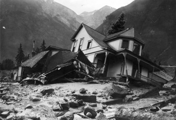 Exterior view of Jack Hawkins residence, on Oak Street, filled with mud and debris from the disastrous Cornet Creek flood on July 27, 1914, Telluride, Colorado; shows two-story wood frame Victorian with front porch and second story bay window tilting on side from flood waters, smaller destroyed structure, and the San Juans in background.
