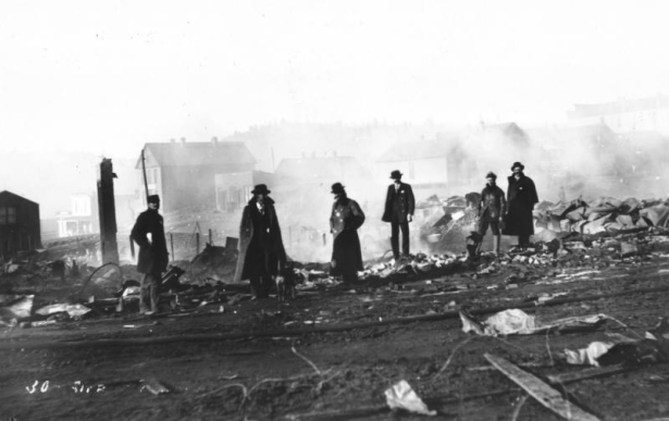 Six men survey destruction from second fire on April 29, 1896, Cripple Creek, Colorado; five men have sherriff badges, some wear overcoats, and one man wears knee-high rubber boots and carries a lunch bucket.