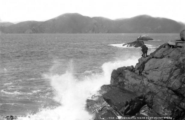 Men stand on rocks as waves splash against the seawall at Fort Point at the Golden Gate channel in San Francisco Bay, San Francisco County, California. Shows the corner of a pillbox.