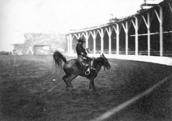 A cowboy rides a bucking horse in a dirt arena during a performance of Buffalo Bill's Wild West Show at Earl's Court in England. The grandstand curves in the right background and is supported by large wood beams.