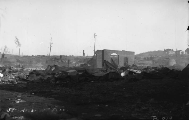 View of desolation caused by second fire on April 29, 1896, Cripple Creek, Colorado; stark remains, twisted sheet metal and debris cover the ground and smoke still rises from ashes; residence or building with hipped roof are in background.