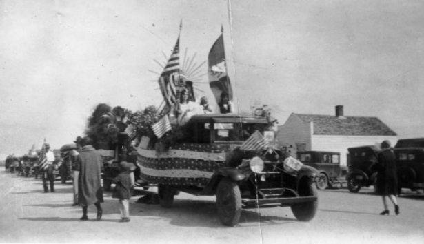View of the annual 16th of September (Diez y Seis de Septiembre, Mexican Independence Day) parade in Sedgwick (Sedgwick County), Colorado. Shows a pickup decorated with bunting, flowers and American and Mexican flags. A woman in a white robe and crown, a woman with braids and possibly a boy in white with a headband sit in front of a sunburst. A line of automobiles is behind the pickup. A man stands near the cars with an American flag.