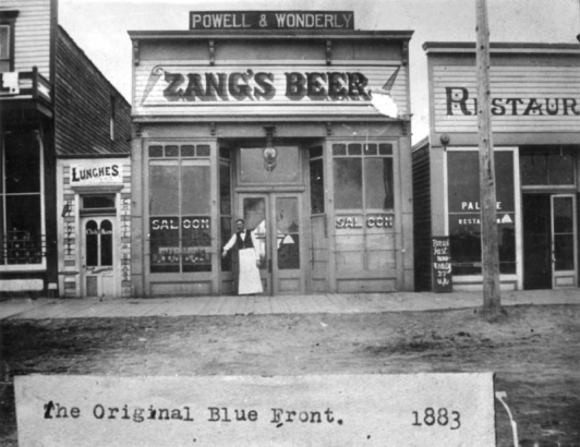 A saloonkeeper wearing an apron stands outside of a saloon in La Junta, Colorado in Otero County.  The nineteenth century, commercial storefront has bracketed cornices, two bays, storefront windows, a transom, and double entry doors.  Bottles of beer are visible in the window.  Signs: "Powell & Wonderly," "Zang's Beer," "Saloon."