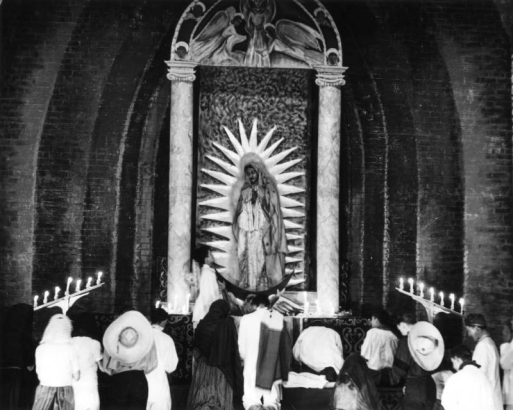 Elementary school children in Greeley, Colorado, in Weld County  reenact the pilgrimage to Guadalupe.  The children are dressed in traditional Mexican costumes, including serapes, sombreros, and mantillas. The children kneel before an altar of Mary.  Horizontal candleabras on both sides of the altar have lit candles.  The altar has two Doric columns and a religious painting above it.