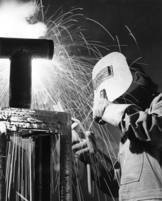 A student welds a metal frame with a threaded t-shaped pipe on the top of it in a welding class in Greeley, Colorado in Weld County.  He holds the welder in his right hand and wears a welding mask and heavy gloves that protect his hands and forearms.