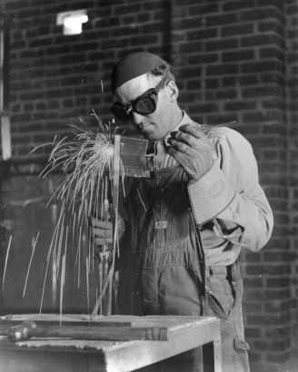 A student welds a piece of metal in a classroom in Greeley, Colorado in Weld County.  He holds the welder in his right hand and the piece of metal in a pair of tongs in his left hand.  He wears a cap, goggles, and heavy work gloves.  He dressed in overalls and a long-sleeved shirt.