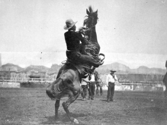 Antonio Esquivel, a champion Mexican vaquero and star of Buffalo Bill's Wild West Show, rides a bucking horse in a dirt arena during a performance of the show at Earl's Court in England. He wears a cowboy hat and a short, dark jacket. A group of cowboys stands in the background watching Esquivel ride. A backdrop with a landscape scene painted on it is at the far end of the arena.