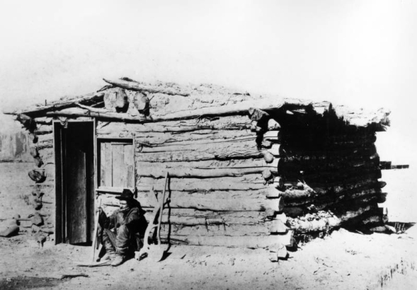 A miner sits outside a crudely built log cabin, Gunnison, Colorado, with his pick ax and shovel. The shack is known as the Sylvester Richardson cabin and the first one in town. The structure has logs of varying sizes chinked with mud, a low sagging front gable roof, hewn log front door and window opening.