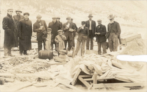 Coal miners, a boy, a teenager, and a young girl stand and eat slices of bread near the remnants of the tent colony destroyed in the Ludlow Massacre during the U.M.W. strike against C.F. & I. in Ludlow (Las Animas County), Colorado. Wood, pieces of canvas, boxes, metal tubs and rubble cover the ground. A suitcase is near the children. The men wear hats, some wear overcoats, the children wear hats and coats.