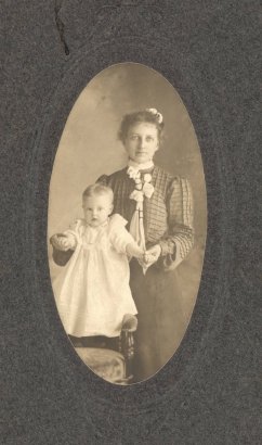 Studio portrait of Jessie Root Thompson and her daughter Helen Elizabeth Thompson. Jessie Thompson wears her hair up; she wears a patterned blouse with a knotted scarf and a dark skirt. Her baby, Helen, wears a white dress. Mrs. Thompson holds her daughter by her hands and balances her on the arm of a chair.