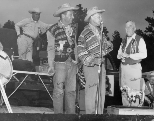 Roundup Riders of the Rockies members stand on a stage during the group's annual trail ride at a camp near the Arapaho Valley Lodge in Grand County, Colorado. Dick Dickson speaks into the microphone. Bob Long wears a straw cowboy hat and smiles. Rudy Paulson wears a woven vest and holds a cigarette. Pete Smythe leans on a piano and stands at the back of the stage. Shows a horse trophy near the microphone.
