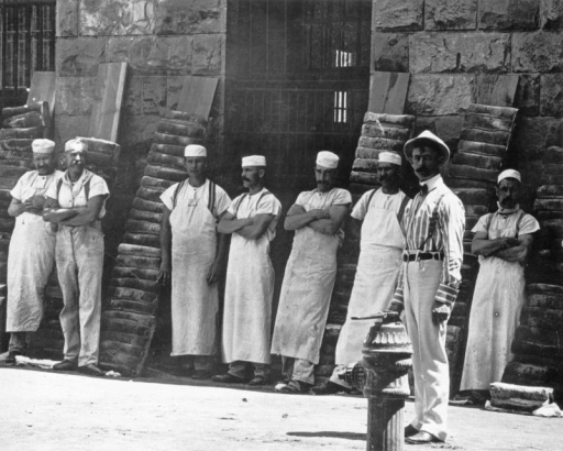 Prisoners in aprons and bakers caps pose near bread stacked against a stone wall of the bakery at the Colorado State Penitentiary in Canon City (Fremont County), Colorado. A man in a striped shirt, a bow tie, suspenders, light pants and a hat poses near the bakers. A fire hydrant is near the men.