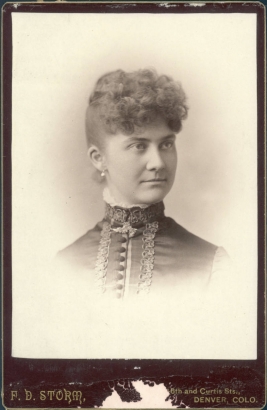 Studio bust portrait of Sallie Bomberger, a senior at Denver High School in Denver, Colorado. Bomberger's bangs are curled and she wears her hair pinned up. Her fitted dress bodice has a band collar, a front button closure, and passementerie trim. She wears pearl drop earrings.