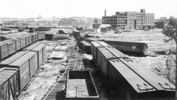 View of the Denver and Rio Grande Western Walker freight yards after the Arkansas River flood in Pueblo, Colorado. Shows twisted and damaged railroad tracks and overturned railroad freight cars. The Atchison, Topeka and Santa Fe bridge, the collapsed D. &.R.G.W. bridge, and the Nuckolls Packing plant are in the distance.
