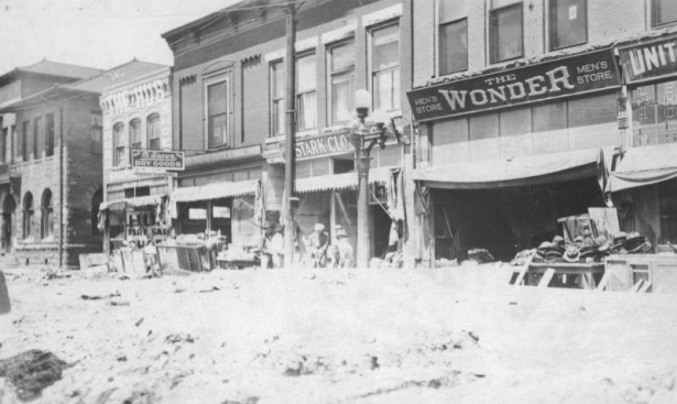 View of flood damaged storefronts on Union Avenue after the Arkansas River flood in Pueblo, Colorado. Men work  with shovels to remove deep mud that washed against the buildings. Shows a utility pole and a lamp post in the street. Hats, clothing and furniture are piled up outside the buildings. Signs on the buildings read: "The Wonder Mens Store," "United Shoes," "Stark Clot[hing Co.]," and "409 M. Braun Dry Goods, Juvenile Clothing."