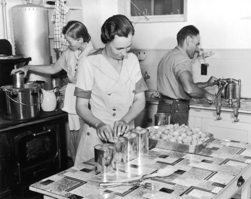A man and two women work in a kitchen canning apricots or peaches for the hot lunch project for the Work Projects Administration in Colorado. One woman stands at a cast iron stove and sterilizes a can in hot water while the other woman stands at a kitchen table and packs fruit into a can. The man seals the can with a hand cranked machine attached to a counter. The table has a linoleum top decorated with an Art Deco pattern.