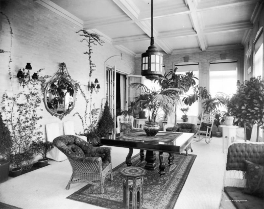 View of a sunroom in the Crawford Hill house at 969 Sherman Avenue, in the Capitol Hill neighborhood of Denver, Colorado.  The room has a tiled floor, brick walls, and a coffered ceiling. A large, square table with a pedestal and four carved legs is in the center of the room. Wicker and slipcovered furniture is scattered throughout the room. A large metal lantern hangs over the table. Shows potted plants , a mirror, and large windows with sheer blinds and shades. An oriental rug is under the table.