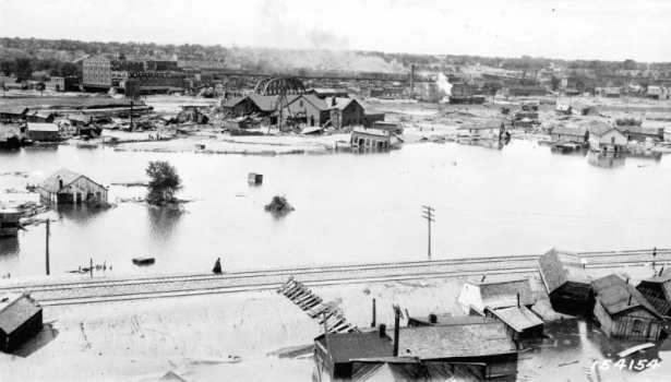 View of flood waters and railroad tracks in Pueblo, Colorado. A woman walks on a railroad embankment; houses, garages, out buildings and tracks are washed up against the embankment. Shows damaged houses and buildings, factories, and a bridge in the flood water and in the distance across the Arkansas River. The Nuckolls Packing Plant is in the distance.