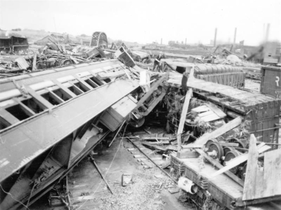 View of an over-turned railroad passenger car, box car and debris after a flood in Pueblo, Colorado. A man picks through the rubble. An intact Atchison, Topeka & Santa Fe bridge, a damaged Denver & Rio Grande Western Bridge, the Arkansas River, and the American Smelting and Refining Company smelter are in the distance.