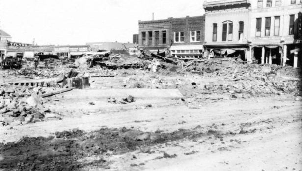 People walk through the debris outside stores destroyed by the Arkansas River flood, on Union Street in Pueblo, Colorado. Signs on commercial buildings read: "Weston's Cafe," "Cut Price Market," "Clothing" and "T. Flynn, Saddles and Harn[esses.]"