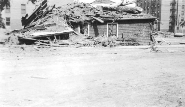 View of a shingle-sided house covered with a tree trunk, mud, and debris after a flood on the Arkansas River in Pueblo, Colorado. Multi-story brick buildings are in the distance.