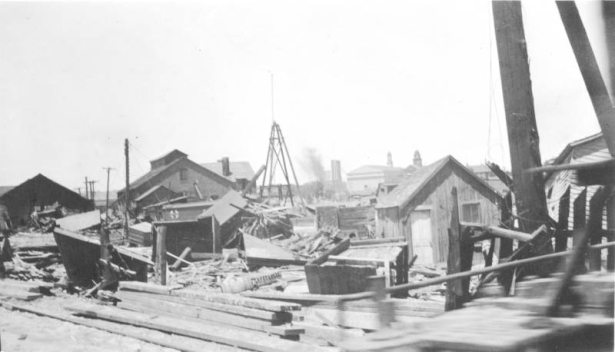View of buildings and shacks moved and destroyed by the flood waters of the Arkansas River in Pueblo, Colorado. Debris, a boxcar, lumber, and utility poles are scattered among the buildings. Smoke stacks and steeples are in the distance.