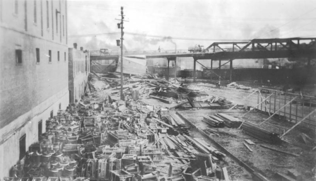 Cast iron cooking stoves are piled up against the side of the brick Holmes Hardware Store at 400 S. Union Avenue near railroad tracks after the Arkansas River flood,  in Pueblo, Colorado. Wreckage and debris are piled up near a viaduct.  Wooden framing braced up with boards is beside the track. Railroad boxcars are in the distance.