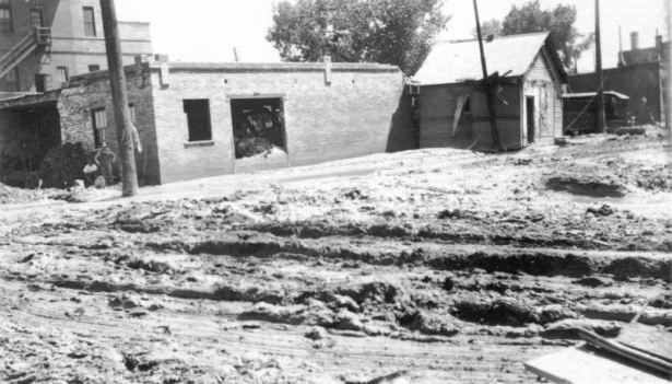 View of a small house that was carried by the flood waters of the Arkansas River and came to rest against the side of a brick garage in Pueblo, Colorado. The garage has damaged walls. Two men pose beside the garage. Ruts in the deep mud are near the buildings.