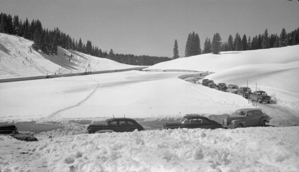 Automobiles are parked along a narrow plowed road at Wolf Creek Pass in Mineral County, Colorado. Shows deep snow, the ski area beside highway US 160, skiers and a rope tow.