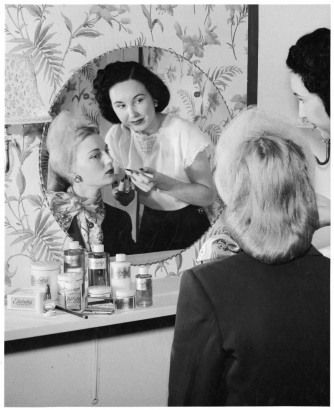 A woman applies lip gloss to a model posed in front of a mirror in probably Denver, Colorado. The model wears veil in her hair and scarf around her neck.  Bottles, jars, and boxes of cosmetics sit on a table, labels read: "Tributa's Studios." Shows floral wallpaper and a sconce.