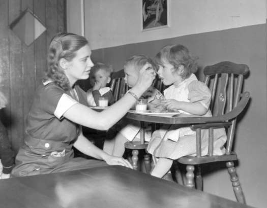 Interior view of toddler eating area, Pueblo Day Care Center (day  nursery), Pueblo, Colorado, shows a woman spoon-feeding three toddlers in high chairs. There are children wearing bibs, and glasses of milk and plates of food on trays.