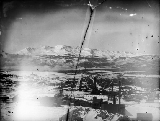 View from Carbonate Hill northwest across Leadville, Colorado, to the south part of town includes mining crushers, smelters with smokestacks, mine tailings, the Midland Railroad's California Gulch Trestle, the Harrison Reduction Works, the Central School, and St. Joseph's. Snow-covered Mount Massive of the continental divide is in the background.