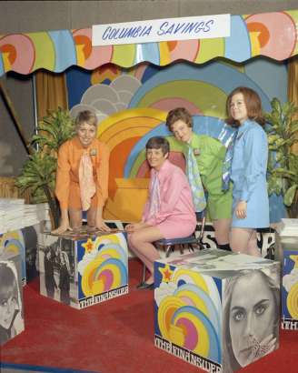 Women pose for a publicity shots near pop-art cubes in Denver, Colorado. A sign on the wall reads: "Columbia Savings," and signs on the cubes read "The Finanswer." The women wear mini-skirts and neckties.