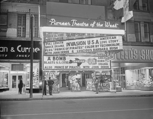 View of the Tabor Theater entrance at 1014 16th (Sixteenth) Street in Denver, Colorado. Signs and marquee read: "Pioneer Theater of the West," "Invasion U.S.A.," "Prince of Pirates," "A Bomb Blasts in U.S. Cities," and "Highlander Blue and Gold Drill Team." Feltman & Curme and Leed's Shoe stores are to the sides.