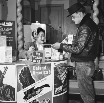 View of a war bonds promotion in a Denver, Colorado theater lobby.  A Japanese woman shows a man a brochure; signs read: "U.S. Bonds Here," "Defend America," and "On Sale Here - Tabor Luck by Caroline Bancroft."
