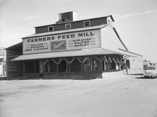 View of "Farmers Feed Mill," at 5870 Wadsworth Boulevard in Denver, Colorado; signs read "Feed Nutrena," and "Rolling Grinding Seed Cleaning Moravian Malting Barley." The building has a pitched monitor roof and a porch.