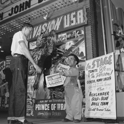 A dwarf hands flyers to a man at the Tabor Theater, 1014 16th (Sixteenth) Street in Denver, Colorado. Signs read: "Invasion U.S.A.," "Prince of Pirates," "A Bomb Blasts in U.S. Cities," and "On Stage Tonite 8:15 p.m. in Person! General Henry Larsen, Chief Civilian Defense Plus Highlander Blue and Gold Drill Team, World Famous Tabor."