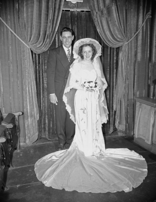 A bride and groom pose for a wedding studio portrait in (probably) Denver, Colorado. The bride's wedding dress has a puddle train; she wears a wide brimmed hat and carries a trailing orchid bouquet. The man wears a suit and tie.